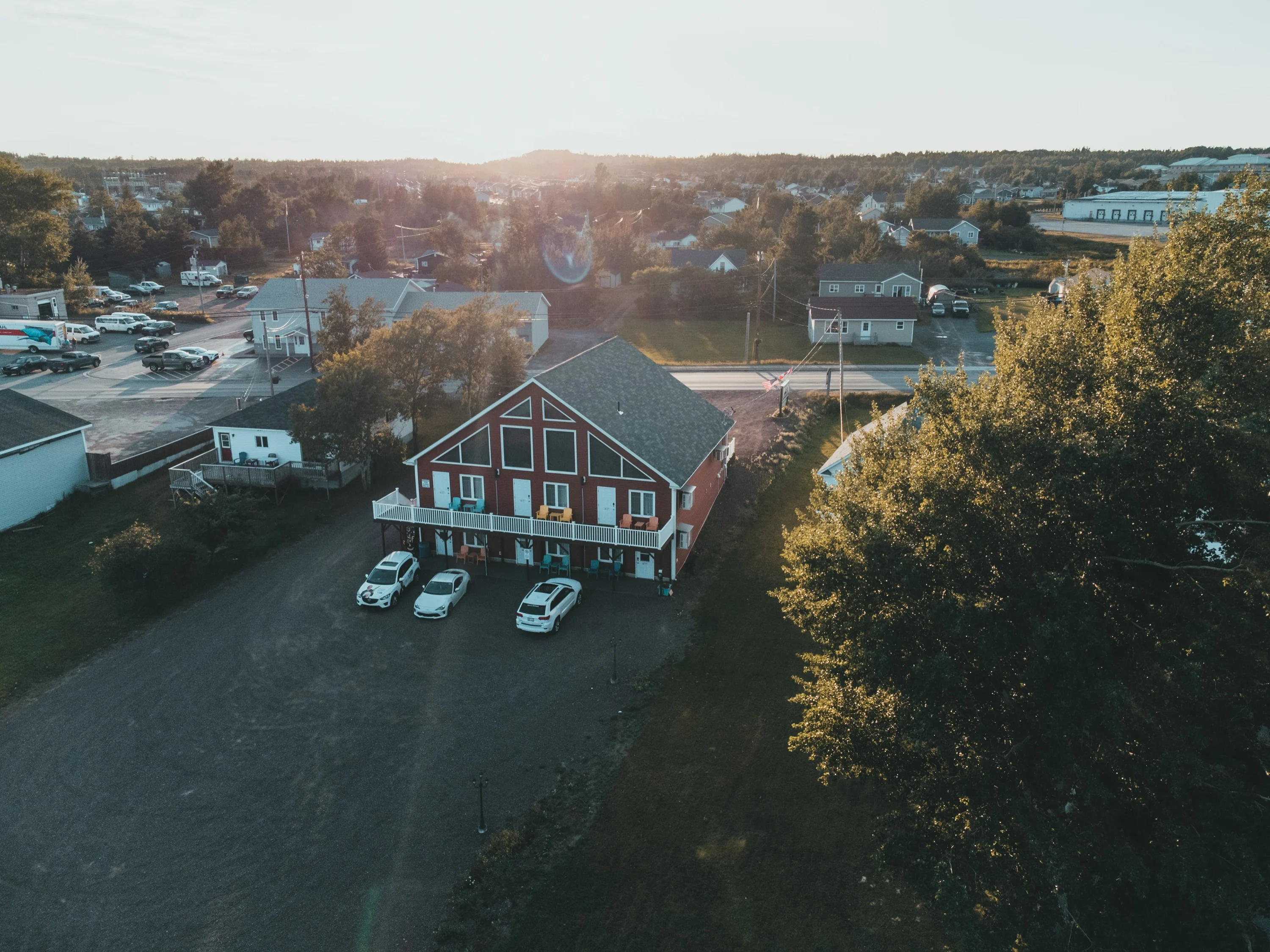 Aerial view at sunset of Islands' Villa, a red chalet-style building with a white balcony and colourful Adirondack chairs, with the town of Lewisporte behind it