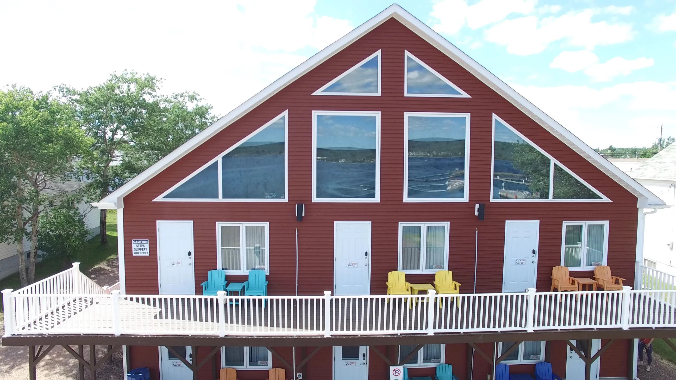The A-frame front of Islands' Villa on a bright day, the bay and sky reflected in its tall triangular windows, above a two-level deck lined with turquoise and yellow chairs