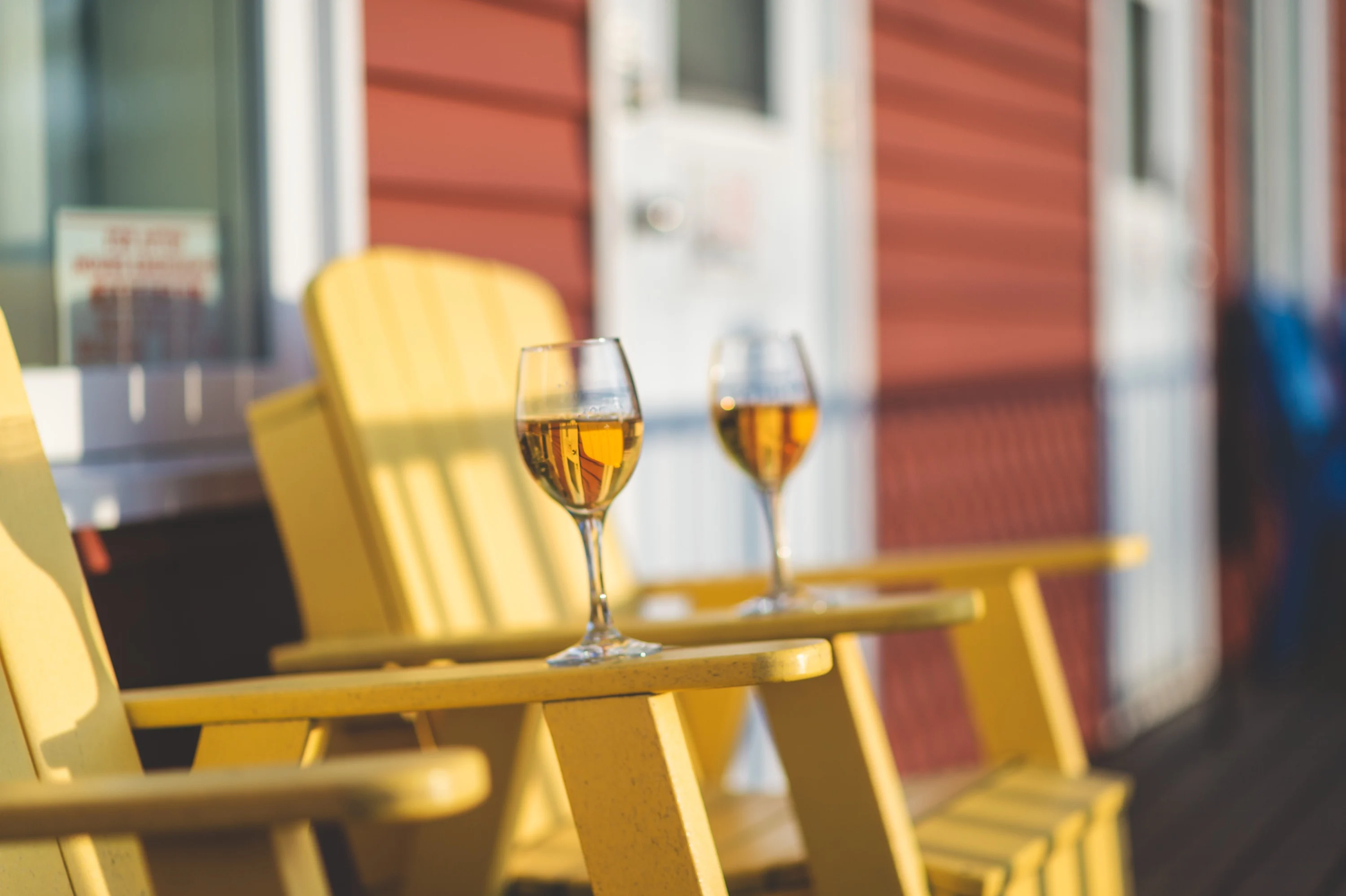 A glass of white wine catching the evening light on the arm of a yellow Adirondack chair, with the red siding of the villa behind